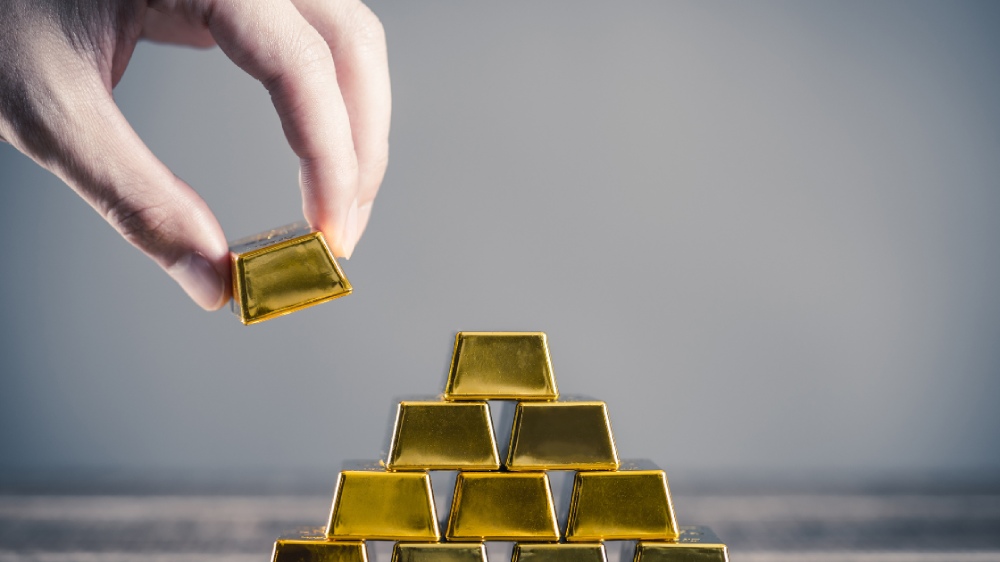 یک دست در حال چیدن آخرین شمش کوچک طلا بر روی هرمی از شمش‌ها