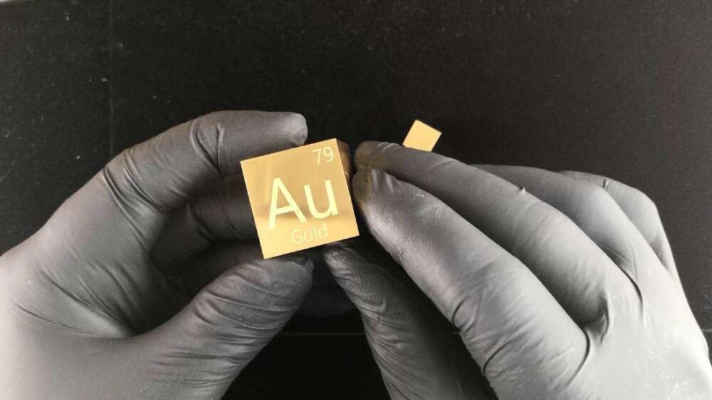 دستهایی با دستکشهای مشکی که مکعبی از عنصر طلا را به همراه اطلاعات جدول تناوبی گرفتهاند