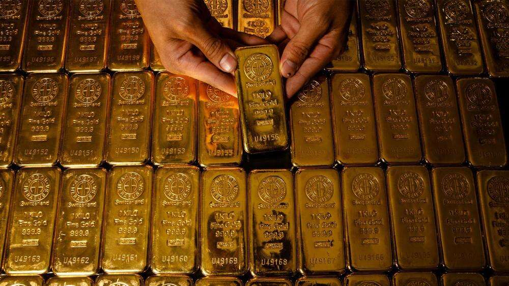 دو دست که یک شمش طلای یک کیلویی را بالای انبوهی از شمشهای طلا نگه داشتهاند