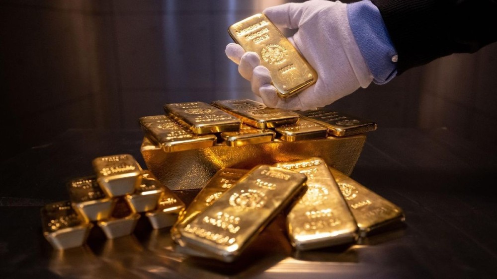 دستی با دستکش سفید در حالی که یک شمش طلا را بالای انبوهی از شمشهای دیگر گرفته است