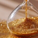 نمای نزدیک از ساعت شنی شیشه‌ای با شن‌های طلایی در حال ریزش